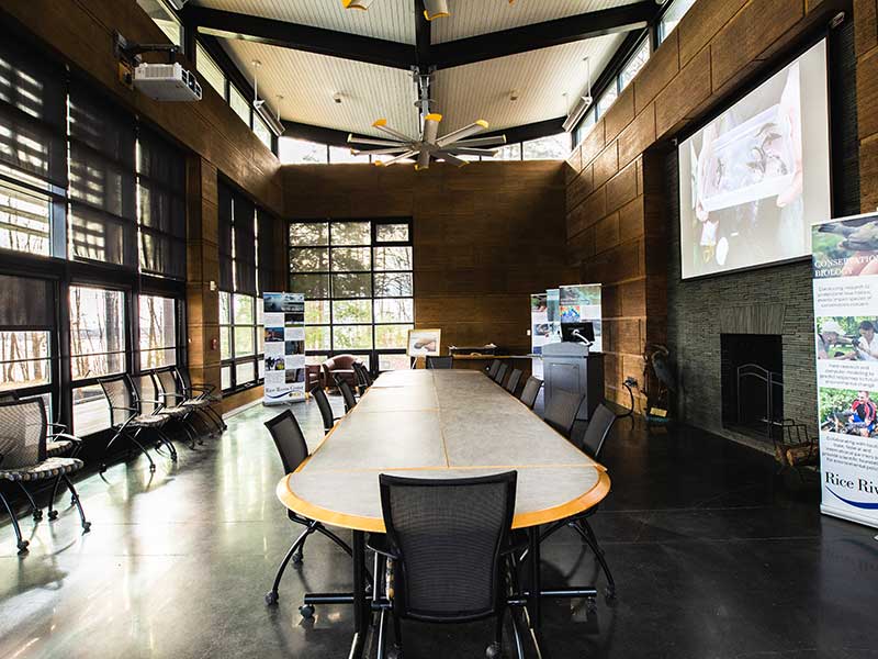 a sunny conference room with a long work table and a large digital screen over a fireplace