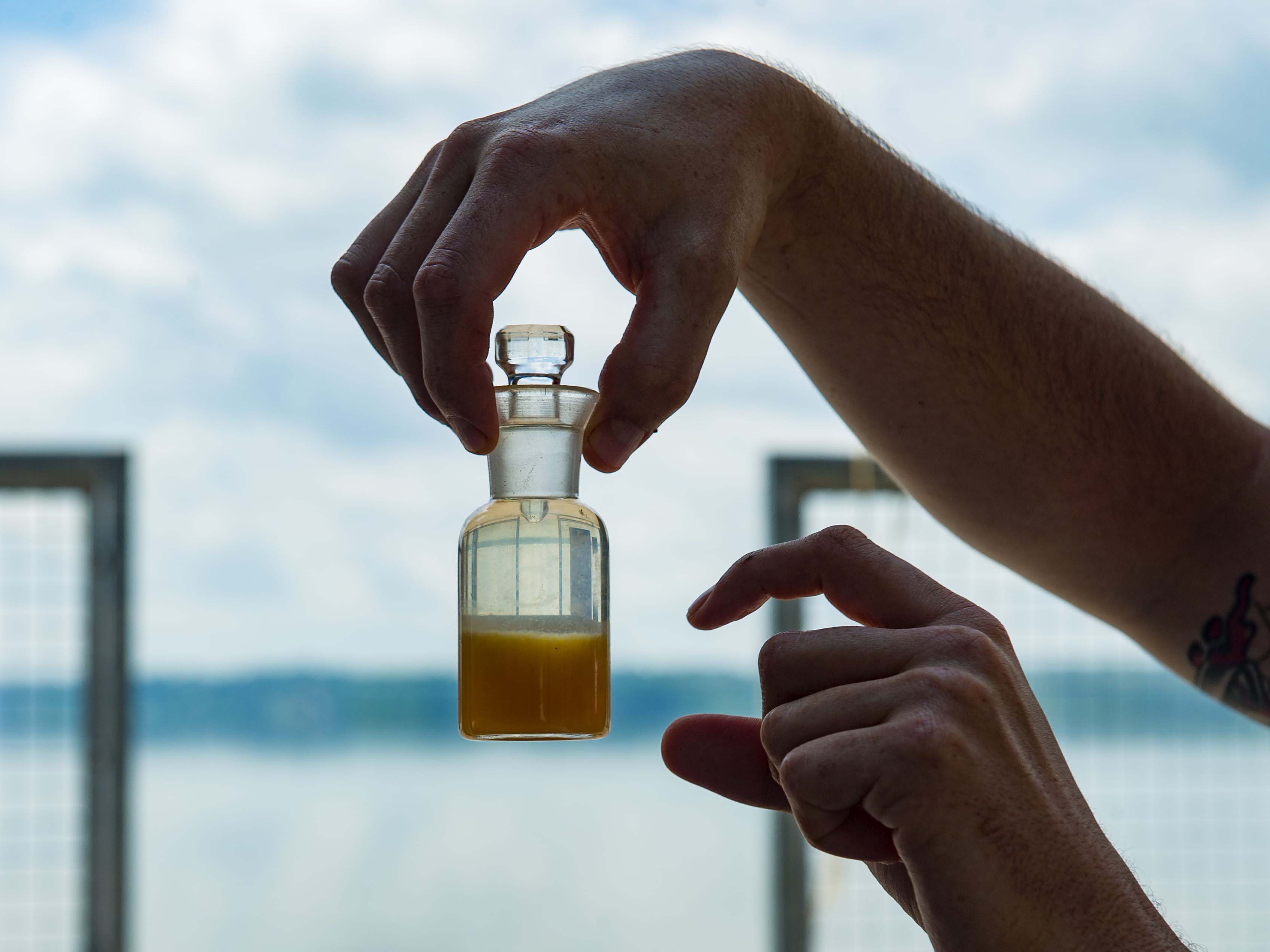 Right holding a vial of murky water, left hand demonstrating amount of water in vial, water in the background